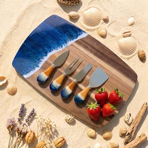 May include: A wooden cheese board with a blue and white wave design on one end. The board includes four cheese tools with orange and blue handles. Fresh strawberries and seashells surround the board, creating a beachy theme.