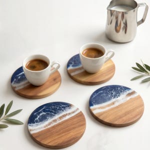 May include: Four round wooden coasters featuring a blue and white resin design that mimics ocean waves. Two white ceramic espresso cups filled with dark coffee sit on two of the coasters, and a stainless steel milk pitcher is nearby.