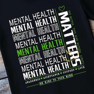 May include: Black t-shirt with the words "MENTAL HEALTH" in white, stacked vertically. One instance of "MENTAL HEALTH" is in green. The word "MATTERS" is in white vertically on the right. The shirt also says "YOU MATTER, YOUR MIND MATTERS, ALWAYS."