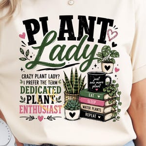 May include: A cream-colored t-shirt featuring a graphic print with the text "PLANT LADY" in black and green, "CRAZY PLANT LADY? I PREFER THE TERM DEDICATED PLANT ENTHUSIAST" in black, green, and pink, and a stack of books labeled "EAT SLEEP WATER PLANTS REPEAT."