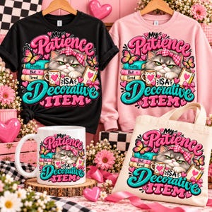 Op de afbeelding: Een zwart t-shirt, een roze sweatshirt, een witte keramische mok en een beige draagtas, elk met een afbeelding van een norse kat met een roze strik, boeken en de tekst My Patience Is A Decorative Item met roze hartjes.