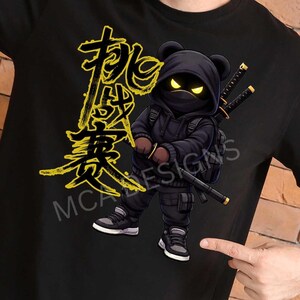 May include: Black t-shirt featuring a cartoon ninja bear with glowing yellow eyes, wearing a black hoodie and holding two katanas. The design includes yellow Japanese characters and the text "MCA DESIGNS".