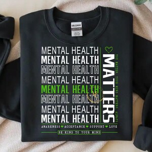 May include: Black sweatshirt with the words "MENTAL HEALTH" stacked vertically in white, with some lines in green. The word "MATTERS" is printed vertically in white with a green heart. The text "YOU MATTER, YOUR MIND MATTERS, ALWAYS" is printed vertically in green. The text "BE KIND TO YOUR MIND" is printed at the bottom.