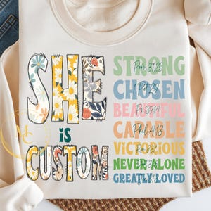 Könnte beinhalten: Cremefarbenes Sweatshirt mit den Worten "SHE IS CUSTOM" in einem floralen Design. Das Wort "SHE" ist in einem hellblauen Blumendesign, "IS" ist in Weiß und "CUSTOM" ist in einem Blumendesign. Zusätzliche Wörter sind "STRONG", "CHOSEN", "BEAUTIFUL", "CAPABLE", "VICTORIOUS", "NEVER ALONE" und "GREATLY LOVED".