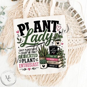 May include: A white canvas tote bag featuring the text "PLANT LADY" in black and green, with additional text "CRAZY PLANT LADY? I PREFER THE TERM DEDICATED PLANT ENTHUSIAST". Includes illustrations of potted plants, a stack of books, and hearts.