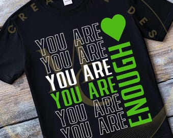 May include: Black t-shirt with the words "YOU ARE" stacked vertically in white outlined text. The word "ENOUGH" is printed vertically in green. A green heart is above the word "ENOUGH".
