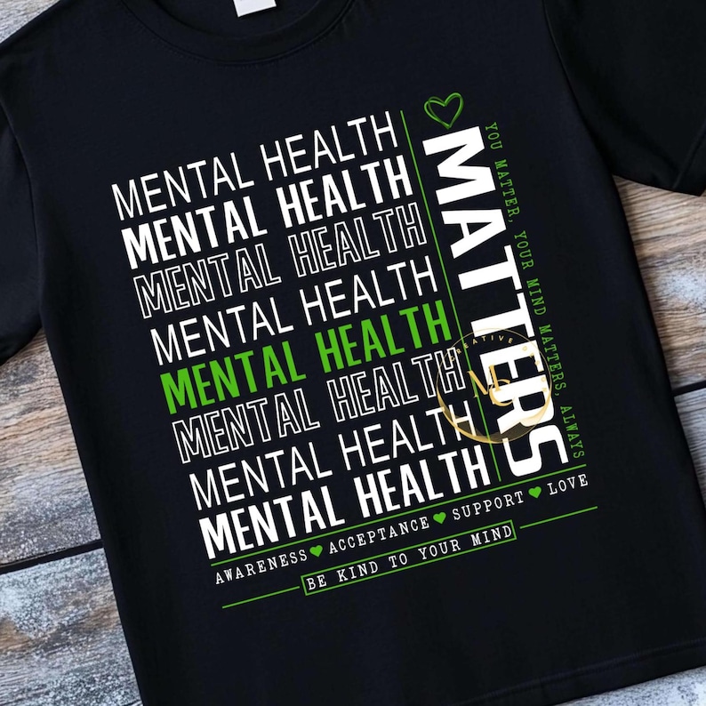 May include: Black t-shirt with the words "MENTAL HEALTH" in white, stacked vertically. One instance of "MENTAL HEALTH" is in green. The word "MATTERS" is in white vertically on the right. The shirt also says "YOU MATTER, YOUR MIND MATTERS, ALWAYS."
