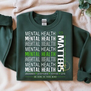 May include: Dark green sweatshirt with the words "MENTAL HEALTH" stacked vertically in white, with one instance in green. The word "MATTERS" is printed vertically on the right side. The phrase "BE KIND TO YOUR MIND" is printed at the bottom.