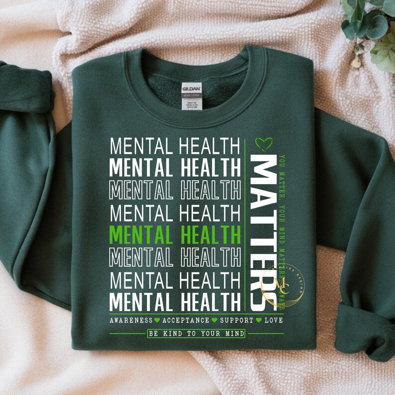 May include: Dark green sweatshirt with the words "MENTAL HEALTH" stacked vertically in white, with one instance in green. The word "MATTERS" is printed vertically on the right side. The phrase "BE KIND TO YOUR MIND" is printed at the bottom.