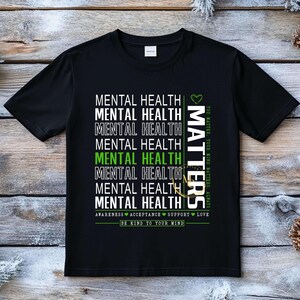 May include: Black t-shirt with the words "MENTAL HEALTH" repeated vertically in white, with one instance in green. The word "MATTERS" is printed vertically in white with a green heart. The shirt also says "BE KIND TO YOUR MIND."