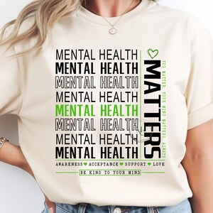May include: Cream-colored t-shirt with the words "MENTAL HEALTH" stacked vertically in black and green. The word "MATTERS" is printed vertically on the right side. The shirt also includes the phrases "AWARENESS ACCEPTANCE SUPPORT LOVE" and "BE KIND TO YOUR MIND."