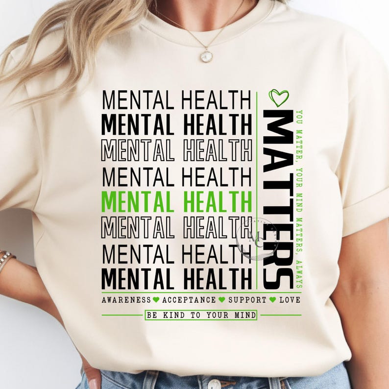 May include: Cream-colored t-shirt with the words "MENTAL HEALTH" stacked vertically in black and green. The word "MATTERS" is printed vertically on the right side. The shirt also includes the phrases "AWARENESS ACCEPTANCE SUPPORT LOVE" and "BE KIND TO YOUR MIND."