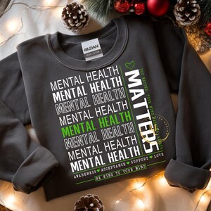 May include: Dark gray sweatshirt with the words "Mental Health" repeated in white, with one instance in green. The word "MATTERS" is printed vertically in white. The sweatshirt also has the text "Awareness, Acceptance, Support, Love" and "Be Kind to Your Mind."