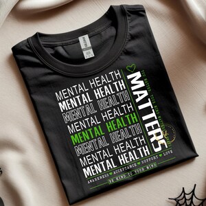 May include: Black t-shirt with the words "MENTAL HEALTH" in white, repeated multiple times. One instance of "MENTAL HEALTH" is in green. The word "MATTERS" is printed vertically in white. Additional text includes "YOU MATTER, YOUR MIND MATTERS", "AWARENESS ACCEPTANCE SUPPORT LOVE", and "BE KIND TO YOUR MIND".