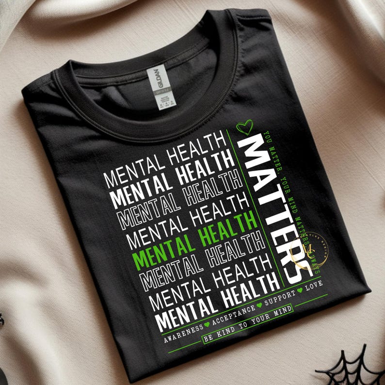 May include: Black t-shirt with the words "MENTAL HEALTH" in white, repeated multiple times. One instance of "MENTAL HEALTH" is in green. The word "MATTERS" is printed vertically in white. Additional text includes "YOU MATTER, YOUR MIND MATTERS", "AWARENESS ACCEPTANCE SUPPORT LOVE", and "BE KIND TO YOUR MIND".