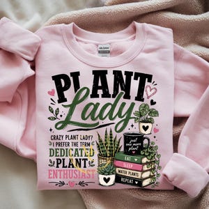 May include: A light pink crewneck sweatshirt featuring a graphic print with the text "PLANT LADY" in black and green, followed by "CRAZY PLANT LADY? I PREFER THE TERM DEDICATED PLANT ENTHUSIAST" and a stack of books labeled "EAT," "SLEEP," "WATER PLANTS," "REPEAT."