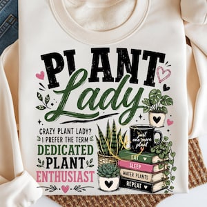 May include: A cream-colored sweatshirt featuring the text "PLANT LADY" in black and green, with additional text "CRAZY PLANT LADY? I PREFER THE TERM DEDICATED PLANT ENTHUSIAST". Graphics include potted plants, a mug, and a stack of books.
