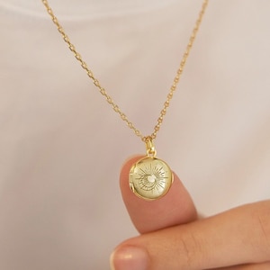 Puede incluir: Un collar dorado con un colgante redondo con un diseño de sol. El colgante cuelga de una cadena delicada. El collar se muestra sobre un fondo blanco.