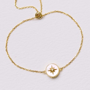 Puede incluir: Pulsera ajustable dorada con un dije circular con diseño de estrella. El dije tiene un fondo blanco y una estrella dorada. La pulsera es delicada y se encuentra sobre una superficie blanca.