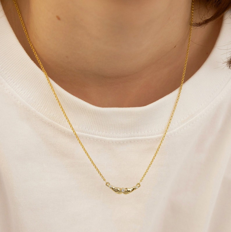 K&ouml;nnte beinhalten: Eine goldene Kettenkette mit einem kleinen Diamanten-Akzent und einem Anh&auml;nger in Form von zwei ineinandergreifenden H&auml;nden.