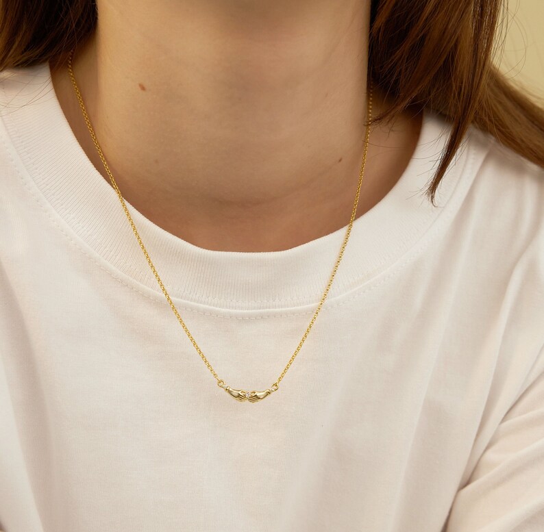 K&ouml;nnte beinhalten: Eine goldene Kettenkette mit einem kleinen, zarten Anh&auml;nger, der zwei ineinandergreifende H&auml;nde zeigt.