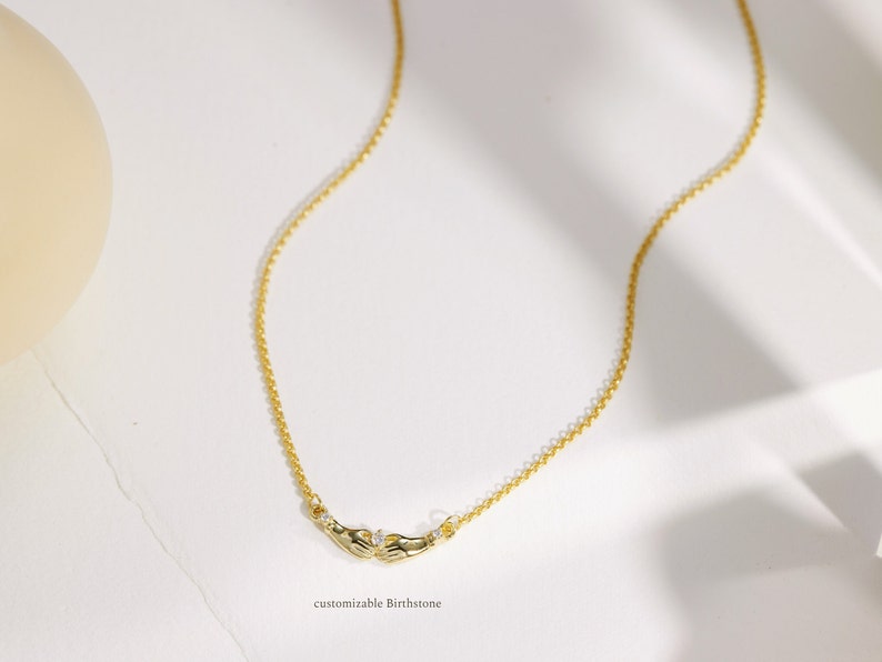 K&ouml;nnte beinhalten: Eine goldene Kettenkette mit einem zarten Design, das zwei H&auml;nde zeigt, die einen kleinen, klaren Stein halten. Die Kette ist mit einem Geburtsstein personalisierbar.