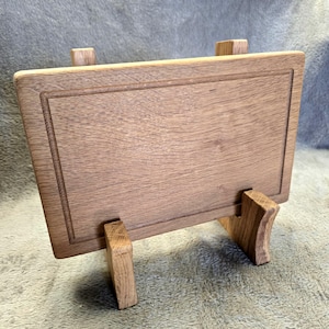 May include: A rectangular wooden chopping board with a visible grain pattern and a recessed border line, supported by two matching wooden stands. The board is light brown in colour and features a smooth, flat surface with a clean, simple design.