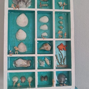 May include: A white frame with a turquoise background holds a collection of seashells and miniature figurines. The frame is on a turquoise easel.