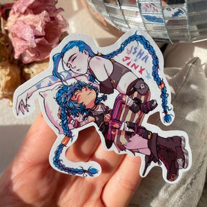 Könnte beinhalten: Ein Aufkleber mit zwei Comicfiguren, eine mit blauen Haaren und einem weißen Kissen, die andere mit blauen Haaren und einem rosa-violetten Outfit. Der Text "Jinx & Isha" steht auf dem Aufkleber.