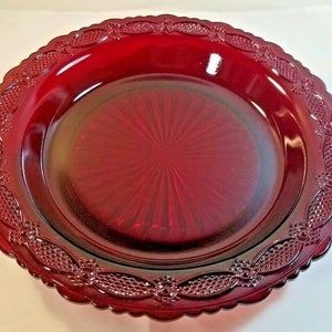 May include: A deep red glass plate with a scalloped edge and a decorative pattern around the rim. The plate has a starburst pattern in the center.