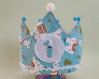 Op de afbeelding: Een turquoise stoffen kroon met een lama-patroon en het nummer 1. De kroon heeft witte kanten randen, blauwe pompons en een witte pompon bovenop. Ontworpen voor een eerste verjaardag.