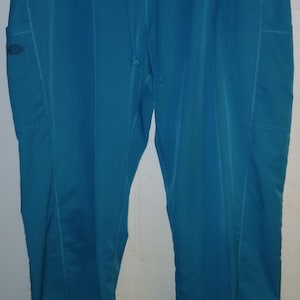 May include: Teal-colored scrub pants with a drawstring waist and two side pockets. The pants are made of a smooth, wrinkle-resistant fabric and hang from a metal hanger. The Dickies logo is visible on the left pocket.