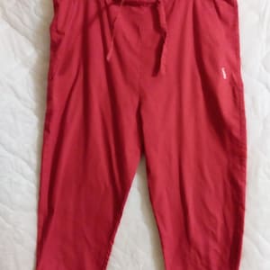 May include: A pair of solid red cropped pants featuring an elasticized waistband with a matching drawstring tie. The pants have a straight-leg cut that ends mid-calf and include a small white logo detail on the upper right side of the garment.