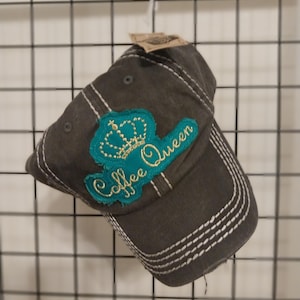 May include: A charcoal gray baseball cap featuring a teal patch with a gold embroidered crown and the text Coffee Queen. The cap has prominent white contrast stitching along the seams and a frayed edge detail on the brim and patch.