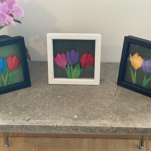 Puede incluir: Tres arreglos de flores de papel origami enmarcados. Cada marco tiene un esquema de color diferente: rosa, azul y amarillo. Los marcos son negros y blancos.