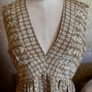 Puede incluir: Vestido sin mangas con cuello en V, exhibido en un maniqu&iacute;. El vestido presenta un estampado geom&eacute;trico y floral en marr&oacute;n y blanco. La tela parece ligera, adecuada para climas c&aacute;lidos. El vestido tiene un corpi&ntilde;o ajustado y una falda fluida.