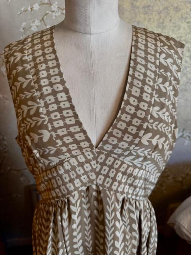 Puede incluir: Vestido sin mangas con cuello en V, exhibido en un maniqu&iacute;. El vestido presenta un estampado geom&eacute;trico y floral en marr&oacute;n y blanco. La tela parece ligera, adecuada para climas c&aacute;lidos. El vestido tiene un corpi&ntilde;o ajustado y una falda fluida.