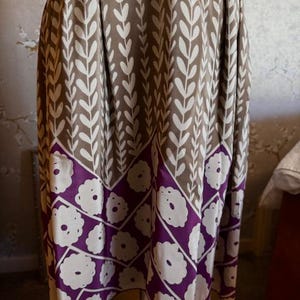 Puede incluir: Un vestido hasta la rodilla con un fondo marr&oacute;n y un estampado de hojas blancas. La parte inferior del vestido presenta una banda morada con dise&ntilde;os de flores blancas. El vestido est&aacute; hecho de una tela fluida.