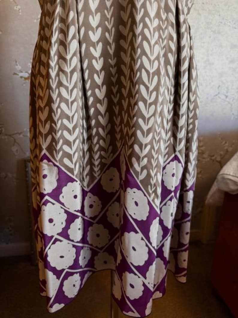 Puede incluir: Un vestido hasta la rodilla con un fondo marr&oacute;n y un estampado de hojas blancas. La parte inferior del vestido presenta una banda morada con dise&ntilde;os de flores blancas. El vestido est&aacute; hecho de una tela fluida.