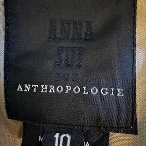 Puede incluir: Una etiqueta negra con el texto "ANNA SUI" y "ANTHROPOLOGIE" en blanco. Debajo, una etiqueta m&aacute;s peque&ntilde;a con el n&uacute;mero "10" en blanco. El fondo es una tela color beige.