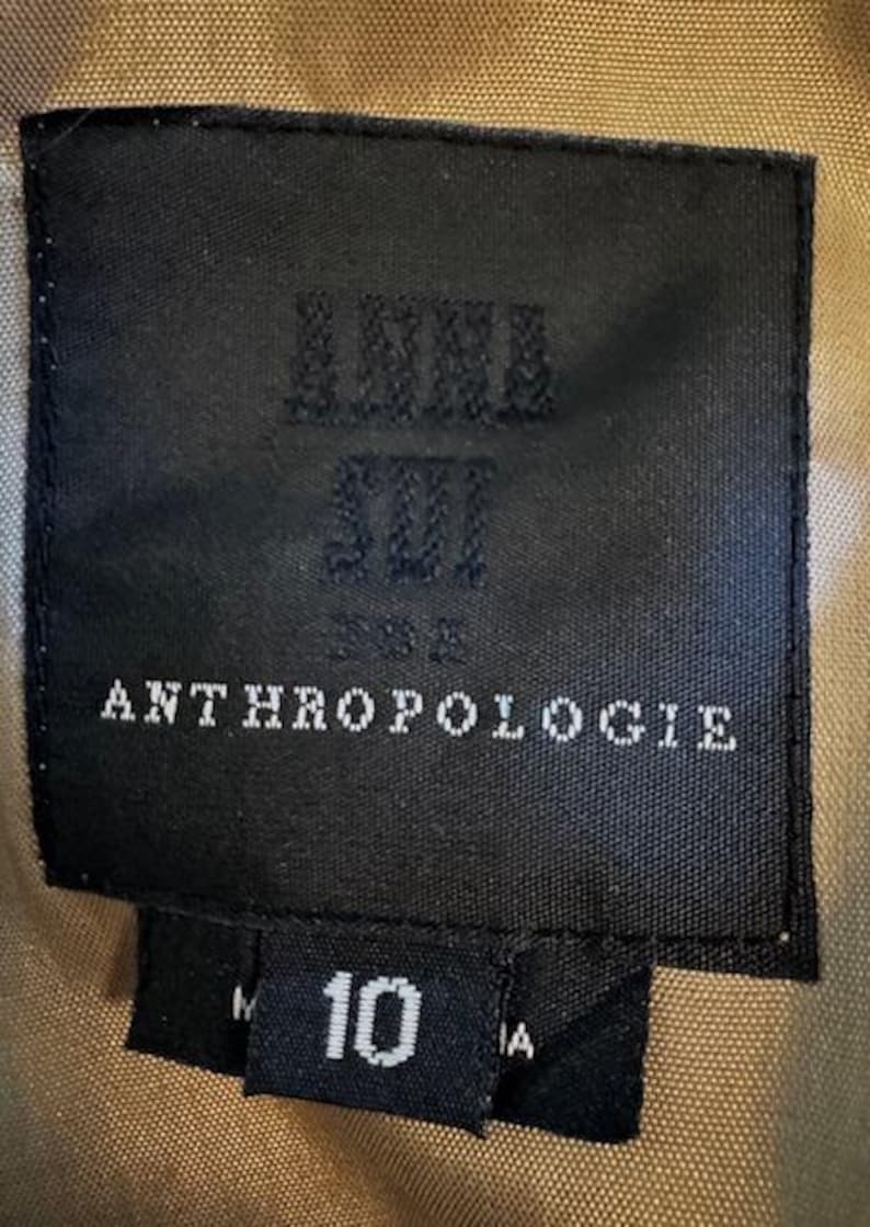 Puede incluir: Una etiqueta negra con el texto "ANNA SUI" y "ANTHROPOLOGIE" en blanco. Debajo, una etiqueta m&aacute;s peque&ntilde;a con el n&uacute;mero "10" en blanco. El fondo es una tela color beige.