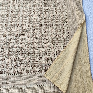 Op de afbeelding: Een beige en bruin textiel met een herhalend geometrisch patroon. De stof is gevouwen en onthult een lichtere beige onderkant. De randen hebben een gedetailleerde rand. Het textiel lijkt een deken of sprei te zijn.