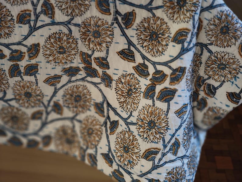 K&ouml;nnte beinhalten: Nahaufnahme eines wei&szlig;en Stoffes mit einem floralen Muster in Braun und Blau. Das Design zeigt gro&szlig;e Blumen und Bl&auml;tter, die durch Stiele verbunden sind. Der Stoff scheint eine Steppdecke oder ein &auml;hnliches Textil zu sein.