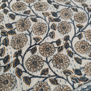 K&ouml;nnte beinhalten: Wei&szlig;e Blumensteppdecke mit einem sich wiederholenden Muster aus stilisierten Blumen und Bl&auml;ttern in Beige- und Blaut&ouml;nen. Das Design ist mit einem kontrastierenden blauen Faden auf den wei&szlig;en Stoff gen&auml;ht, wodurch ein struktureller Effekt entsteht. Die Steppdecke ist gefaltet.