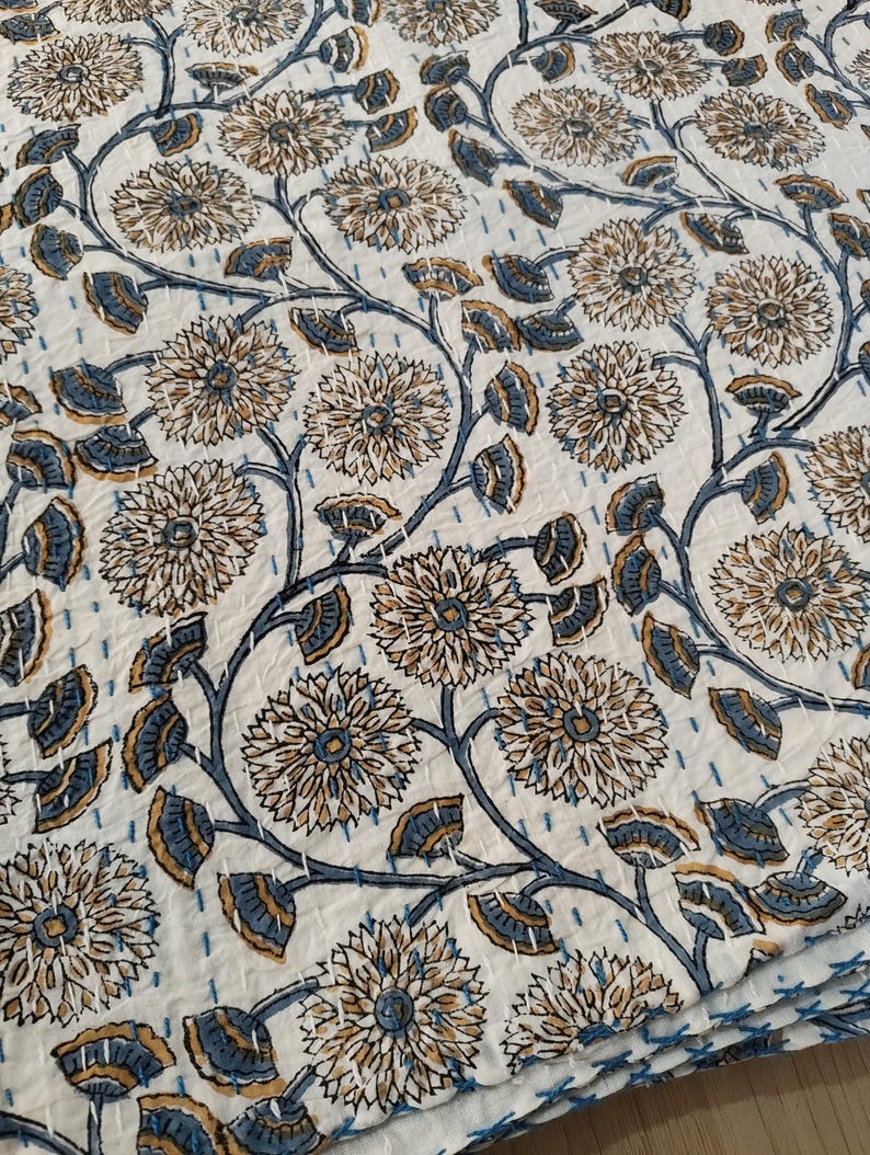 K&ouml;nnte beinhalten: Wei&szlig;e Blumensteppdecke mit einem sich wiederholenden Muster aus stilisierten Blumen und Bl&auml;ttern in Beige- und Blaut&ouml;nen. Das Design ist mit einem kontrastierenden blauen Faden auf den wei&szlig;en Stoff gen&auml;ht, wodurch ein struktureller Effekt entsteht. Die Steppdecke ist gefaltet.