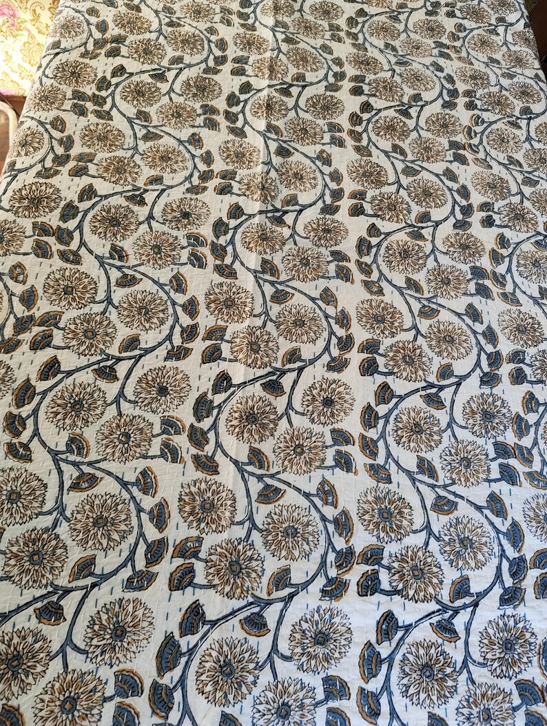 K&ouml;nnte beinhalten: Eine cremefarbene Blumensteppdecke mit einem sich wiederholenden Muster aus braunen und blauen Blumen und Ranken. Die Steppdecke hat ein durchgehendes Stichdesign, das Textur und Details hinzuf&uuml;gt. Das Gesamtdesign ist ein klassisches Blumenmuster.