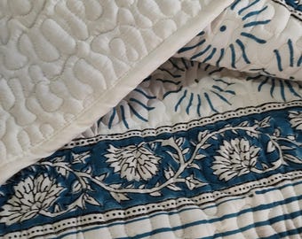 May include: A quilted bedspread featuring a white top layer with a wavy stitched texture. The folded corner reveals a white base fabric decorated with blue floral patterns, intricate vine motifs, and horizontal blue stripes on a white background.