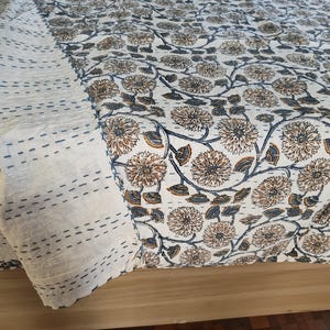 K&ouml;nnte beinhalten: Eine cremefarbene Steppdecke mit einem Blumenmuster in Braun- und Blaut&ouml;nen. Die Steppdecke hat einen gen&auml;hten Rand und ist &uuml;ber einer Holzoberfl&auml;che drapiert. Das florale Design umfasst detaillierte Blumen- und Blattmotive.
