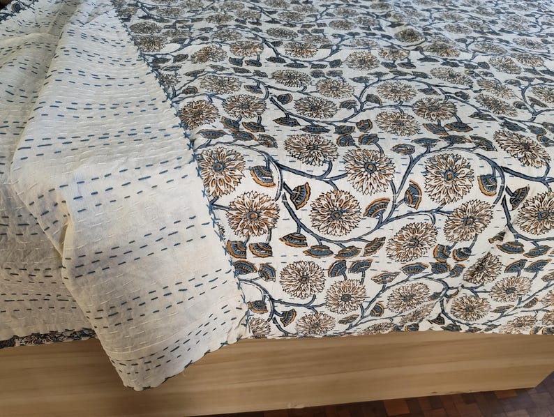 K&ouml;nnte beinhalten: Eine cremefarbene Steppdecke mit einem Blumenmuster in Braun- und Blaut&ouml;nen. Die Steppdecke hat einen gen&auml;hten Rand und ist &uuml;ber einer Holzoberfl&auml;che drapiert. Das florale Design umfasst detaillierte Blumen- und Blattmotive.