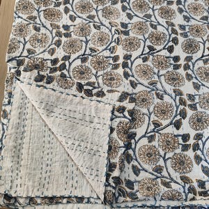 K&ouml;nnte beinhalten: Eine gefaltete wei&szlig;e Steppdecke mit einem floralen Muster in Braun- und Blaut&ouml;nen. Die Steppdecke weist ein sich wiederholendes Design aus stilisierten Blumen und Ranken auf. Die Kanten sind mit blauen N&auml;hten versehen, und die Steppdecke ist gefaltet, um die Unterseite freizulegen.